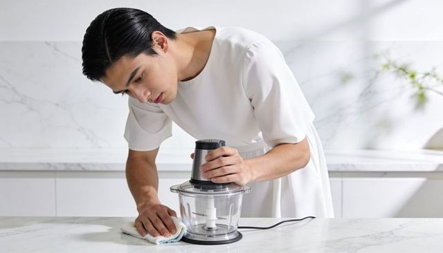 Leaning man wearing white short-sleeve dress cleaning food processor on marble countertop