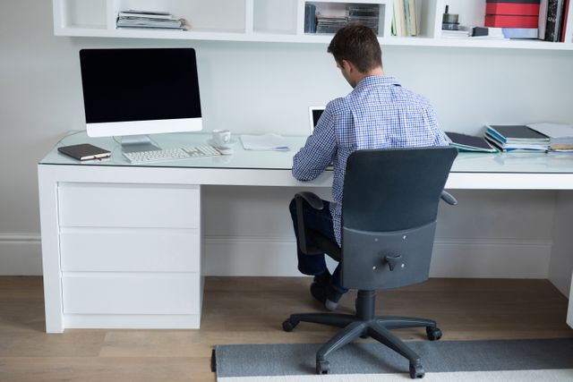 Man sitting at desk in modern home office, working on computer. Ideal for illustrating remote work, home office setups, productivity, and professional work environments.