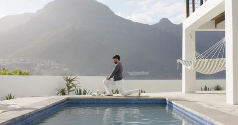 Indian man practicing yoga by modern pool with mountain view and hammock on minimal terrace