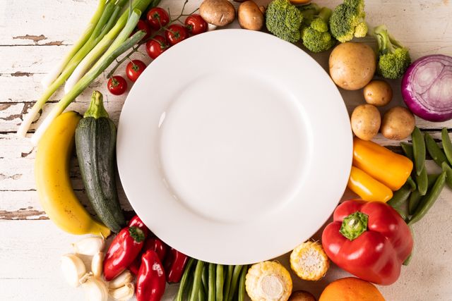 Empty white plate surrounded by an assortment of fresh vegetables including bell peppers, tomatoes, broccoli, potatoes, zucchini, and more. Ideal for use in healthy eating, organic food, and nutrition-related content. Perfect for blogs, websites, and advertisements promoting vegetarian or vegan diets, cooking recipes, and farm-to-table concepts.