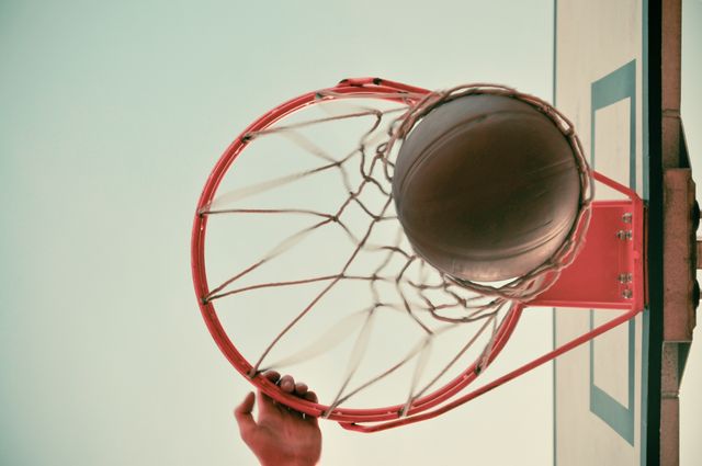 Basketball going through hoop from below view with a player's hand visible. Concept for sports achievement, fitness goals, recreational activity, and athletic performance. Ideal for promoting sporting events, fitness programs, and motivational content.