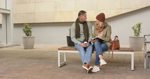 Senior couple sitting on bench in urban plaza wearing scarves holding coffee and notebook