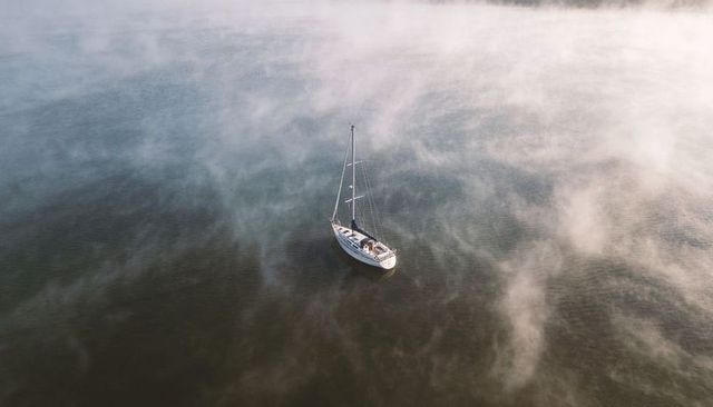 Solitary sailboat drifting through dawn mist over glassy water, peaceful nautical solitude