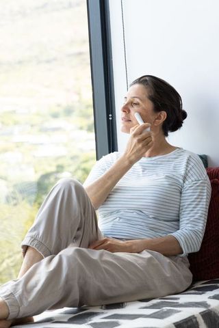 Middle-aged woman relaxing at window seat using handheld facial device for skincare