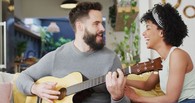 Image of happy diverse couple relaxing in living room, man playing guitar and signing to woman. Happiness, inclusivity, free time, romance, music, togetherness and domestic life.