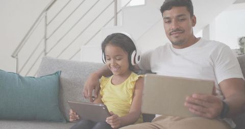 Father sharing tablet time with daughter on couch, headphones, smiling, digital learning