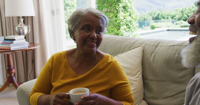 Senior woman holding a cup of coffee, smiling and engaging in conversation while sitting on a comfortable couch in a cozy living room. Ideal for representing themes of relaxation, companionship, and retirement lifestyle. Useful for advertisements, blogs, or articles focusing on senior health, leisure activities, and family bonding.