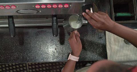 African american female barista frothing milk with steam wand at espresso machine overhead