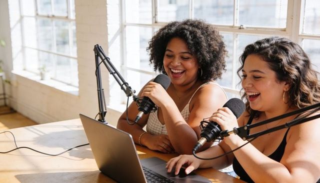 Two women recording lively podcast in sunlit loft studio with laptop and microphones