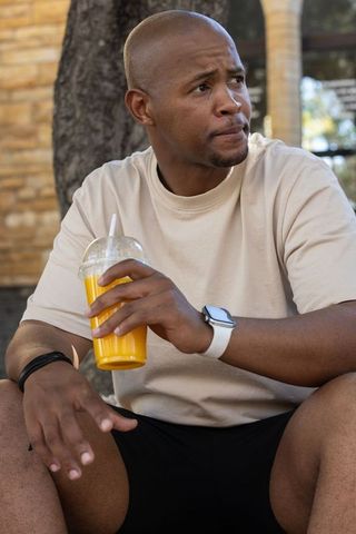 Contemplative african man sitting on bench holding orange juice cup, looking right in courtyard