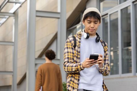 Asian student walking on campus wearing beanie and headphones checking smartphone