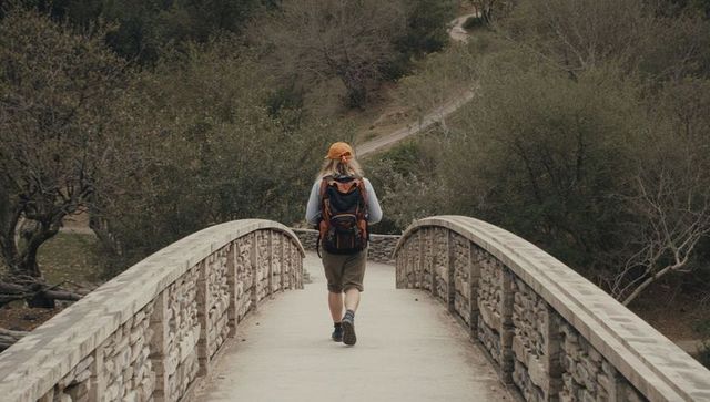 Solo hiker crossing stone bridge on forest trail with orange cap and backpack