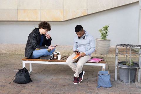 Two male students using smartphones on campus bench studying outdoors with notebooks