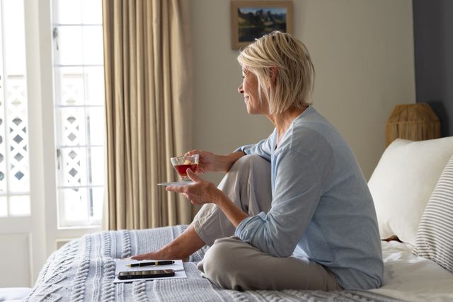 Smiling mature caucasian woman sitting on bed drinking tea and looking out of window, copy space. Domestic life, living alone and senior lifestyle concept.