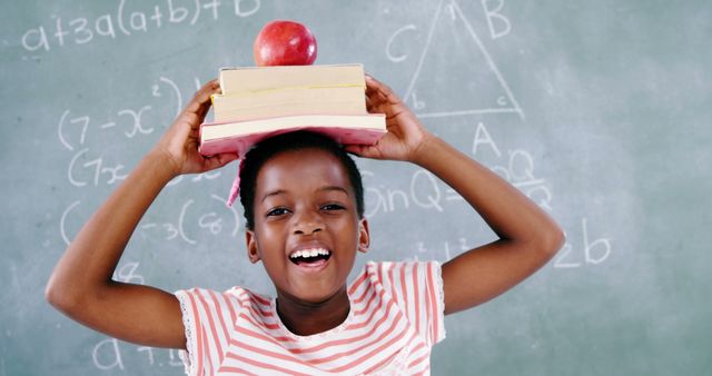 A young African American girl smiles joyfully while balancing books and an apple on her head in front of a chalkboard with mathematical equations, with copy space. Her playful approach to learning highlights the importance of making education engaging and fun for children.
