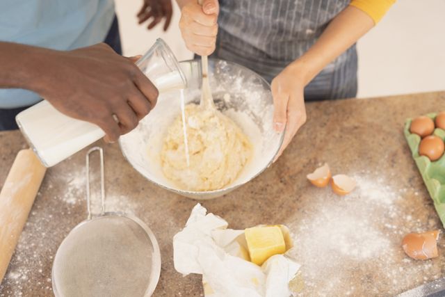 Diverse couple mixing ingredients in a bowl, enjoying a fun and engaging culinary activity. Butter, eggs, and flour are scattered around, emphasizing the homemade aspect of their endeavor. Ideal for use in lifestyle blogs, cooking websites, or advertisements promoting home kitchen products and family bonding activities.