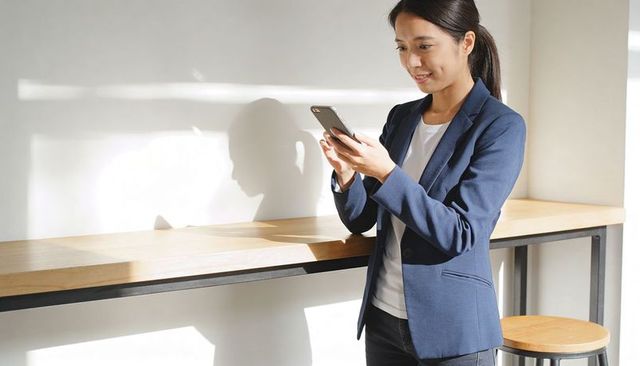 Asian businesswoman checking smartphone by bright coworking counter wearing navy blazer