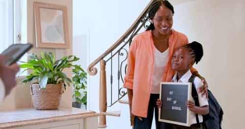 Caption captures mother and daughter smiling at front door holding school sign. Image can be used to represent parenting, childhood milestones, back-to-school moments, and family unity. Suitable for education websites, parenting blogs, or school-related campaigns.
