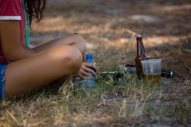 This image shows a woman sitting on the grass during an outdoor festival, with bottles and a plastic cup nearby. Ideal for use in articles or advertisements related to summer festivals, outdoor events, leisure activities, or lifestyle blogs.
