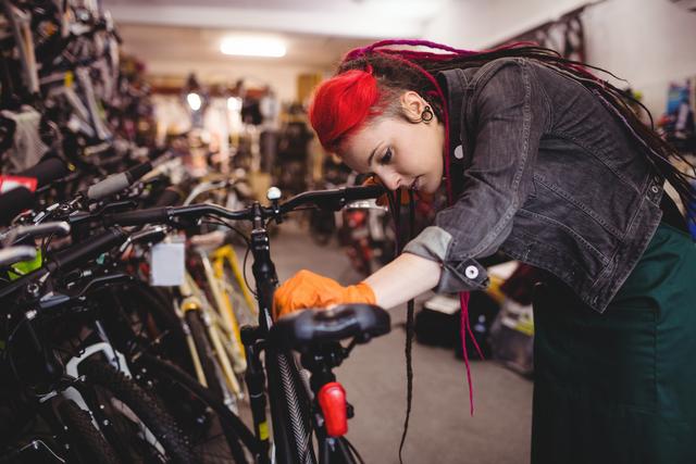 Female mechanic with red hair and dreadlocks examining a bicycle in a workshop. She is wearing a denim jacket and orange gloves, focusing on the bike's details. This image can be used for promoting bicycle repair services, illustrating professional mechanics at work, or highlighting active and sustainable lifestyles.