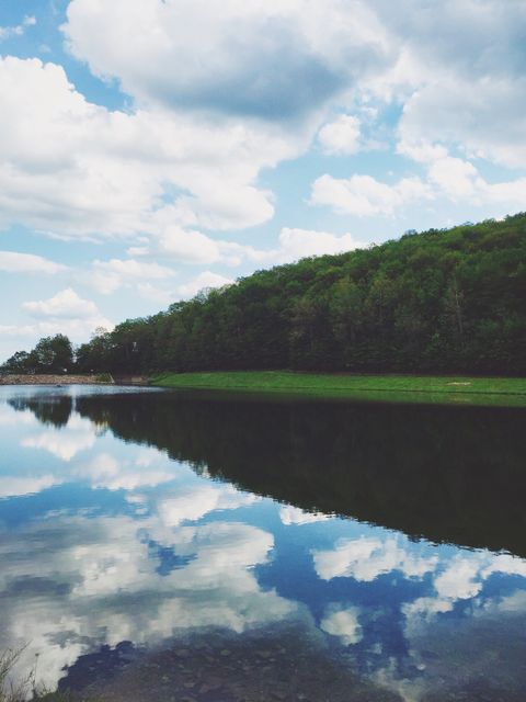 Scenic view of tranquil lake reflecting fluffy clouds and dense green forest. Ideal for projects related to nature, outdoor activities, relaxation, travel, and environmental conservation.