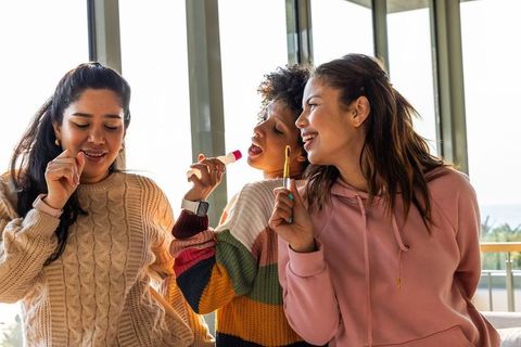 Multicultural friends applying lip balm and laughing on sunlit balcony in cozy sweaters