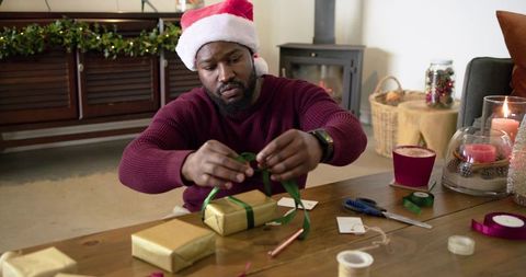 African American man wearing Santa hat wrapping holiday gifts with green ribbon and gold paper