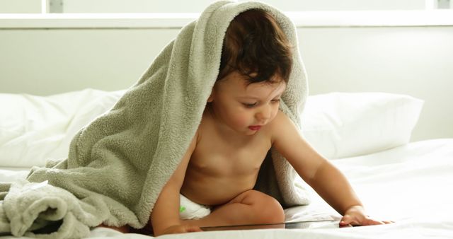 Cute baby with blanket on head playing with tablet on a bed