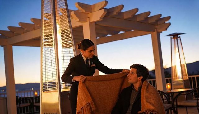 Hotel server wrapping blanket around guest on rooftop terrace at twilight, outdoor heaters