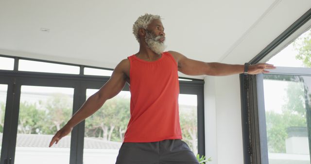 Senior man in red tank top practicing yoga near window at home offering inspiration for healthy living in older age. Ideal for fitness, wellness, senior health care, and home-based workout content.