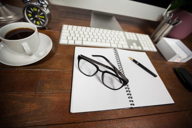 Ideal for illustrating a productive work environment, this image features a spiral notepad, a cup of black coffee, and spectacles on a wooden desk. Perfect for articles or blogs about productivity, office setups, or morning routines. Can also be used in advertisements for office supplies or workspace organization.