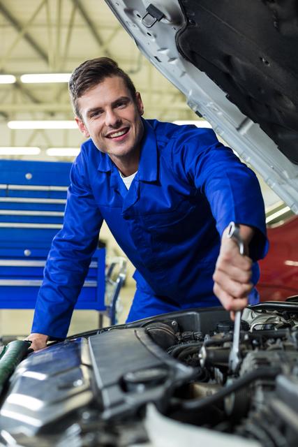 Mechanic in blue uniform smiling while servicing a car engine in a well-lit repair garage. Ideal for use in automotive service advertisements, repair shop promotions, and articles about car maintenance and professional mechanics.