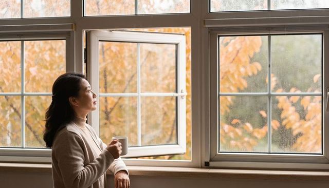 Gazing woman holding mug by rain-streaked window with warm autumn foliage outside