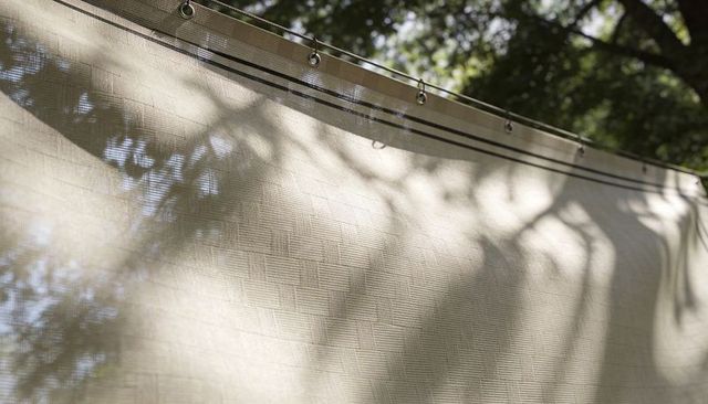 Sunlit beige woven patio shade with metal grommets, wire trim and dappled tree shadows