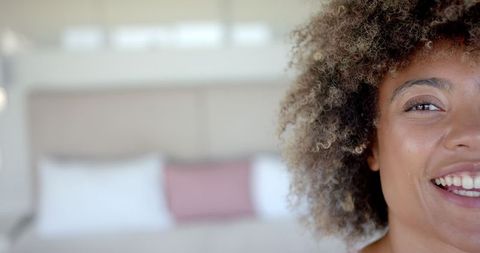 image captures close-up of a smiling woman with curly hair, radiating happiness and contentment inside a cozy home environment. the soft background enhances her joyful, relaxed expression. ideal for use in content associated with positivity, lifestyle guides, home living inspiration, and promoting mental well-being.