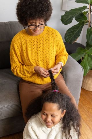 African american woman braiding child's hair on sofa wearing yellow sweater and glasses