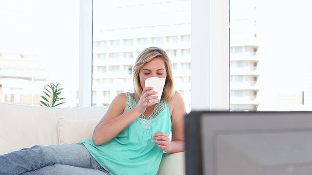 Blonde woman sitting on a white sofa in a bright living room, casually drinking coffee while watching television. The large windows provide ample natural light, creating an airy and comfortable atmosphere. Useful for themes related to home lifestyle, morning routines, comfort, leisure, and relaxation.