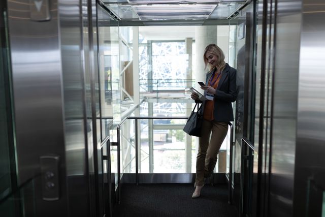 Front view of happy young blonde Caucasian businesswoman using mobile phone in modern office elevator