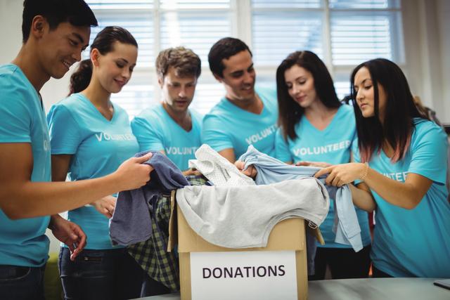 Group of young adults wearing blue volunteer shirts sorting through donated clothes in a community center. Ideal for illustrating concepts of charity, teamwork, community service, and social responsibility. Suitable for use in articles, blogs, and promotional materials related to volunteering, nonprofit organizations, and humanitarian efforts.