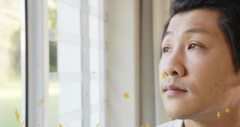 Contemplative asian man gazing out window with gold star stickers, serene indoor portrait