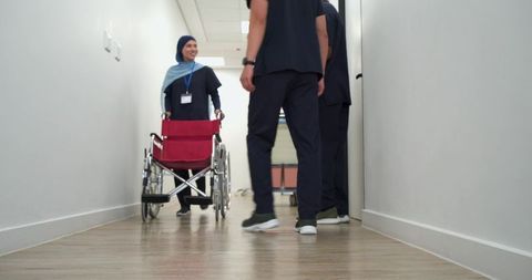 Nurse wearing hijab pushing red wheelchair smiling in healthcare corridor