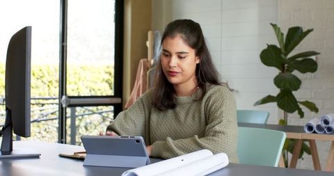 Asian woman working on tablet at desk reviewing blueprints with monitor in home workspace