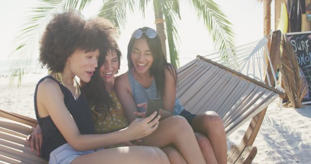 Diverse women sitting on hammock, laughing and looking at smartphone. Summer, free time, chill, vacation, happy time, friendship.