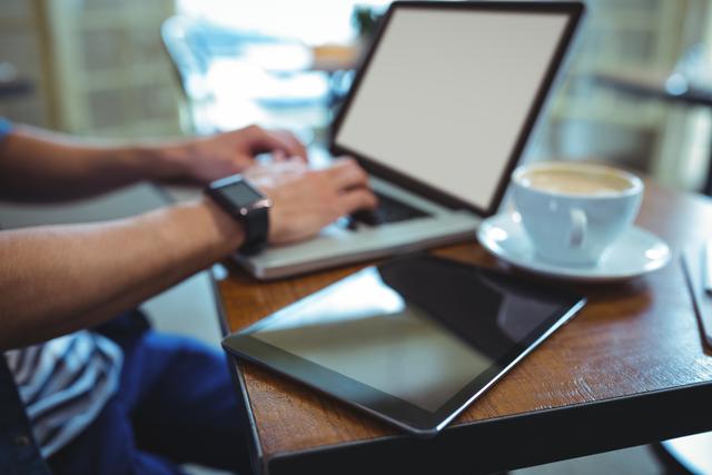 Man working on laptop in a café with a digital tablet and cup of coffee on the table. Ideal for illustrating remote work, freelancing, modern business environments, and digital technology usage in casual settings.