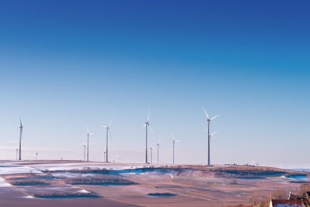 Several wind turbines stand tall on a sprawling field under a clear blue sky. The landscape exhibits a perfect blend of agriculture and advanced green technology, fostering a picturesque view of renewable energy production. This image is ideal for topics on sustainable energy practices, technological advancements in clean power, or environmental conservation initiatives.
