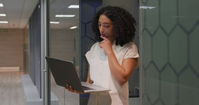 Young African American businesswoman in modern office corridor, thoughtfully analyzing data on a laptop. Ideal for corporate, business, and technology themes, illustrating professionalism, concentration, and advanced work environments.