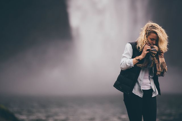 Blonde woman wearing a black vest and scarf standing near a misty waterfall. Perfect for travel blogs, nature exploration themes, and promoting outdoor adventures.
