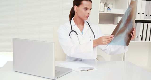 Female doctor carefully analyzing an X-ray in a modern office equipped with a laptop and clipboard. Ideal for content relating to medical diagnostics, healthcare services, hospital promotions, medical training or educational material about radiology.