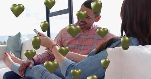Couple sharing smartphone and laughing on coastal sofa, relaxing with floating heart overlay