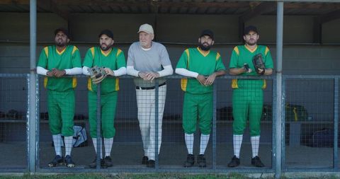 Baseball team leaning on dugout railing with senior coach, players in green uniforms